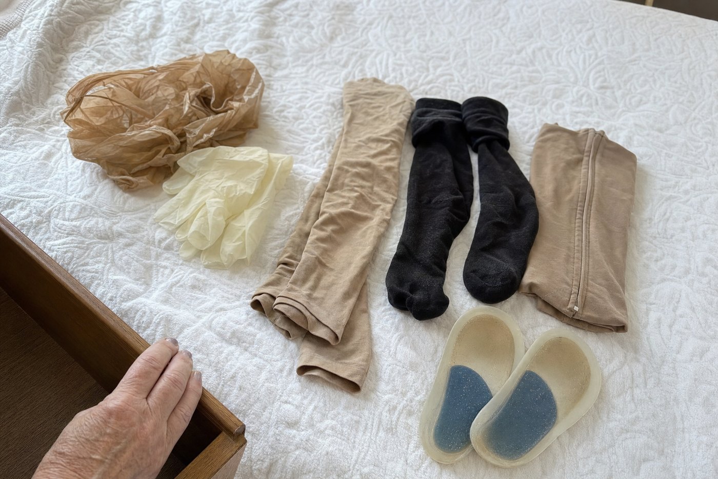 Beige prescription stockings, donning gloves, a plastic bag, rolled-down knee-highs and gel insoles on a bed beside an open drawer